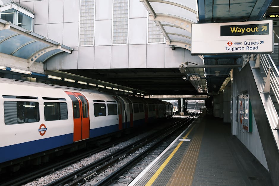 An underground train platform at Hammersmith station, featuring a modern train with a white body, orange and blue accents, and large windows. The platform surface is made of concrete and appears clean, with yellow tactile paving along the edge for safety. Overhead, a curved glass and metal canopy provides shelter, and a bright yellow and black directional sign indicates the way out towards buses and Talgath Road, hanging above the platform. To the right, a staircase with metal handrails leads up to the street level. The station's exterior includes glass and metal panels, reflecting natural light, and the area appears well-maintained and tidy, highlighting the importance of surface cleaning and maintenance in public transport environments. Hammersmith Cleaners emphasizes the need for regular sanitisation and deep cleaning of such surfaces to ensure hygiene and safety.