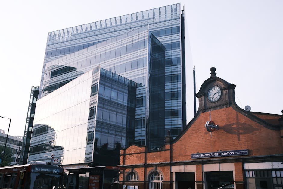 A modern glass office building with reflective windows and sleek exterior, positioned next to the historic brick Hammersmith Station building featuring a classic clock and arched windows, under a clear sky. The scene highlights the contrast between contemporary architecture and traditional railway station design, emphasizing clean surfaces and well-maintained structures. Hammersmith Cleaners specializes in surface cleaning, deep cleaning, and sanitisation services for both residential and commercial clients, ensuring hygiene and spotless environments in areas like offices, homes, and stations.