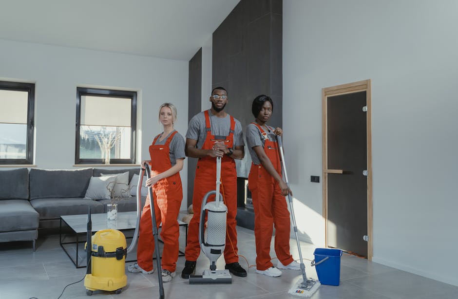 Three professional cleaners from Hammersmith Cleaners are engaged in surface cleaning and deep cleaning tasks inside a modern residential living room. They are dressed in grey shirts and bright orange overalls, standing on a tiled floor with a glossy ceramic finish, which appears clean and free of dust. The group is equipped with various cleaning tools: one with a yellow vacuum cleaner, another with a handheld spray bottle and microfiber cloth, and the third holding a mop with a blue bucket nearby. The room features large windows allowing natural light to illuminate the grey sofa with white and grey cushions and a glass coffee table, emphasizing a tidy and well-maintained space. The walls are painted in neutral tones, with a dark accent wall behind the cleaners. This scene highlights quality domestic cleaning services, focusing on sanitisation and surface maintenance, as part of the services offered by Hammersmith Cleaners to address booking delays and common problems with cleaners in W6, Hammersmith.
