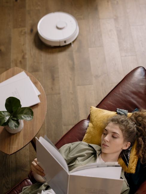A young woman with curly hair and a headband is lying on a brown leather sofa, reading a book with a neutral expression. She is positioned near a yellow cushion, with her head resting on it. To her left, there is a wooden side table holding a small potted plant with broad green leaves and some white papers or notebooks. The room has a wooden floor with a visible robotic vacuum cleaner moving across it, indicating a clean and well-maintained surface. Bright, natural lighting illuminates the area, emphasizing the tidy and hygienic condition of the space, which reflects typical domestic cleaning and surface maintenance practices. The scene relates to routine hygiene and cleanliness, consistent with the services offered by Hammersmith Cleaners, especially in the context of addressing booking delays or common cleaning issues in residential environments.