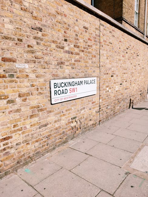 Close-up of a brick wall on Fulham Palace Road in Hammersmith, featuring a white street sign with black text reading 'BUCKINGHAM PALACE ROAD SW1' and a red 'SW1' postcode. The sidewalk in front consists of large concrete slabs, with a small section of green paint visible on one slab. The scene is illuminated by natural daylight, highlighting the textured, weathered bricks and the clean, well-maintained appearance of the area. Hammersmith Cleaners offers surface cleaning and deep cleaning services for residential and commercial properties, ensuring hygiene and presentation are maintained at this level of urban environment.