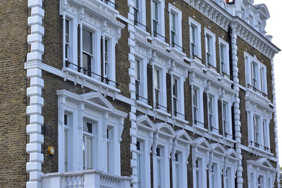 The exterior facade of a multi-story residential building on Fulham Palace Road features detailed white decorative cornices, window frames, and moldings contrasting against the brown brick walls. The building has several large sash windows, each with black metal railings on small balconies, and is well-maintained with clean, polished surfaces. Bright natural light illuminates the building, highlighting the intricate architectural details and pristine condition of the painted trim. The overall appearance suggests a period property, possibly requiring routine cleaning and maintenance. This image is associated with Hammersmith Cleaners' commercial and residential surface cleaning services, including deep cleaning and sanitisation, as featured on their website https://hammersmithcleaners.org.uk.