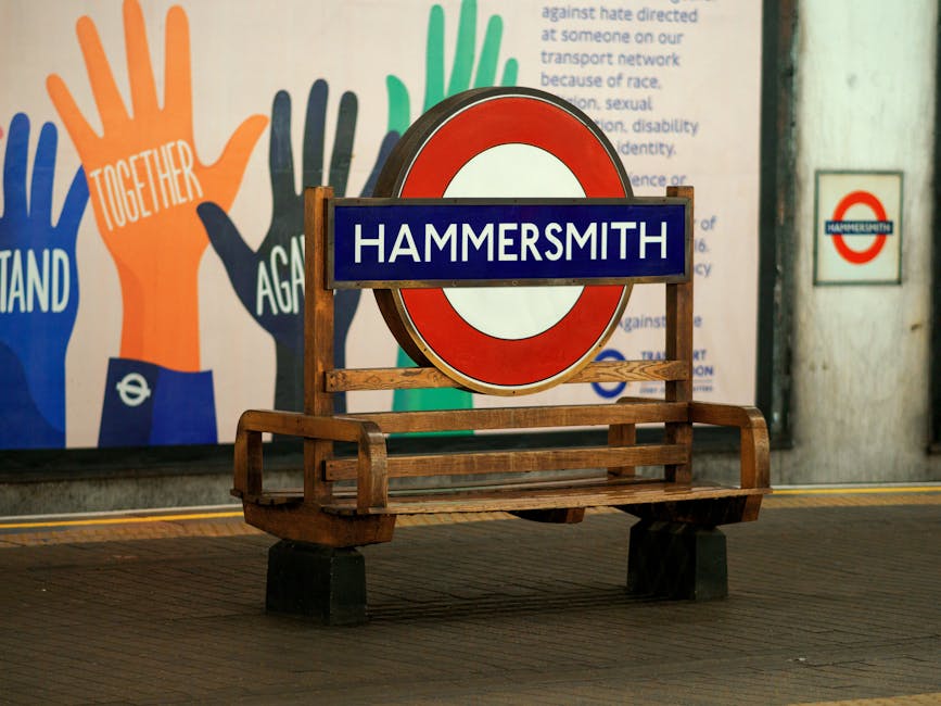 A wooden bench positioned beneath a circular Hammersmith underground station sign, featuring a blue background with white text. Behind the bench, a colorful mural of hands in orange, green, blue, and black, with words like 'together' and 'stand' visible, is painted on the wall. The station sign is mounted on a stand and the surrounding area appears clean and well-maintained, with bright lighting illuminating the scene. To the right, a small Hammersmith logo is attached to the wall. The environment suggests a public transportation setting in Hammersmith, and the image relates to the importance of surface cleaning and maintenance in busy transit areas, with Hammersmith Cleaners providing professional cleaning services.