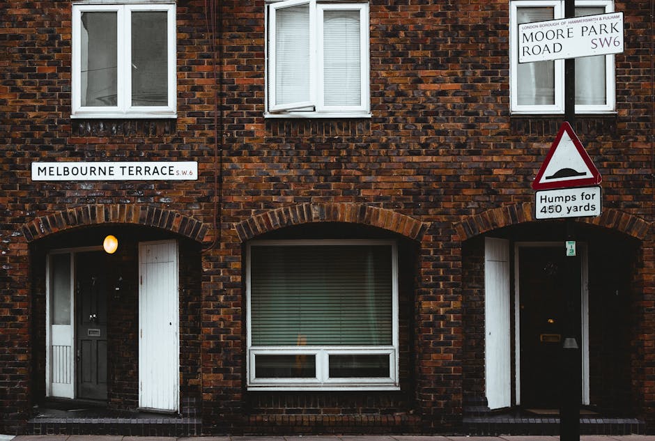 Photograph of a brick residential building on Moore Park Road in Hammersmith, featuring two white-framed double-glazed windows on the upper floor and two arched doorways on the ground floor, one with a white wooden door and the other with a dark door. There are street signs indicating Melbourne Terrace SW6 and Moore Park Road SW6, along with a traffic sign warning of bumps for 450 yards. The building's façade shows weathered red brickwork, and the ground-level windows have closed blinds, with no visible interior clutter or dust, indicating exterior maintenance. The lighting is natural, suggesting daytime, with a single wall-mounted light near the door on the left. This setting exemplifies the need for deep cleaning and surface sanitisation of residential exteriors, an area where Hammersmith Cleaners can assist with domestic cleaning and maintenance services.