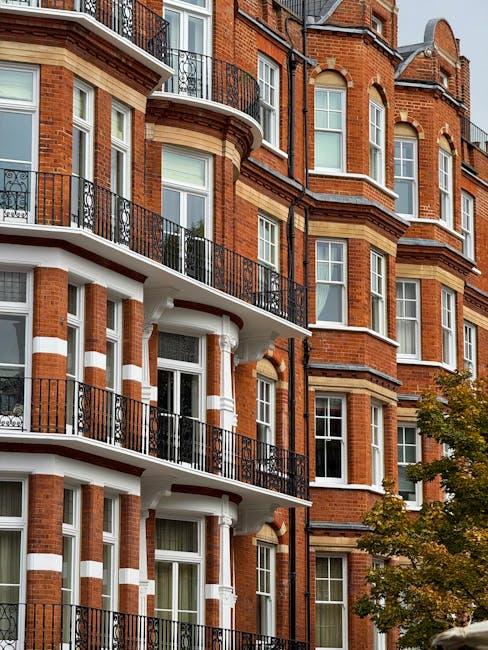 View of a multi-storey red brick residential building with white decorative banding and black wrought iron balconies. The building features large white-framed sash windows, some with glass panes reflecting light, and rounded bay windows with ornate detailing. A tree with green and yellow leaves partially obscures the lower right corner, indicating autumn season. The scene is well-lit with natural daylight, highlighting the clean and well-maintained surfaces of the building's facade. This image exemplifies the architectural style commonly found in Hammersmith, and visually conveys the importance of surface cleaning and maintenance for such historic residential properties, as performed by Hammersmith Cleaners in their deep cleaning and domestic cleaning services for flats in W6, HAMMERSMITH.