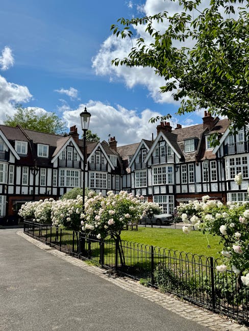 View of a residential street at Ravenscourt Park in W6, Hammersmith, featuring a row of traditional multi-story Tudor-style houses with white walls and black timber framing. The scene includes a well-maintained green lawn bordered by a black metal fence and clusters of blooming white roses along the sidewalk. A classic black lamppost stands near the sidewalk, with a backdrop of lush green trees and a bright blue sky with scattered clouds. The image emphasizes cleanliness and meticulous outdoor maintenance, reflecting professional deep cleaning and landscaping efforts, as seen through the pristine condition of the flower beds and paved walkway. Hammersmith Cleaners specializes in comprehensive cleaning, including surface cleaning and sanitisation of residential exteriors as depicted.