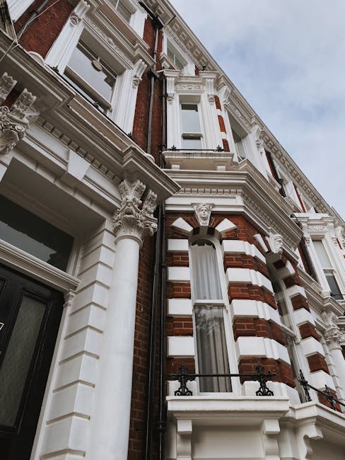 View of a multi-storey red brick residential building with white decorative banding and black wrought iron balconies. The building features large white-framed sash windows, some with glass panes reflecting light, and rounded bay windows with ornate detailing. A tree with green and yellow leaves partially obscures the lower right corner, indicating autumn season. The scene is well-lit with natural daylight, highlighting the clean and well-maintained surfaces of the building's facade. This image exemplifies the architectural style commonly found in Hammersmith, and visually conveys the importance of surface cleaning and maintenance for such historic residential properties, as performed by Hammersmith Cleaners in their deep cleaning and domestic cleaning services for flats in W6, HAMMERSMITH.