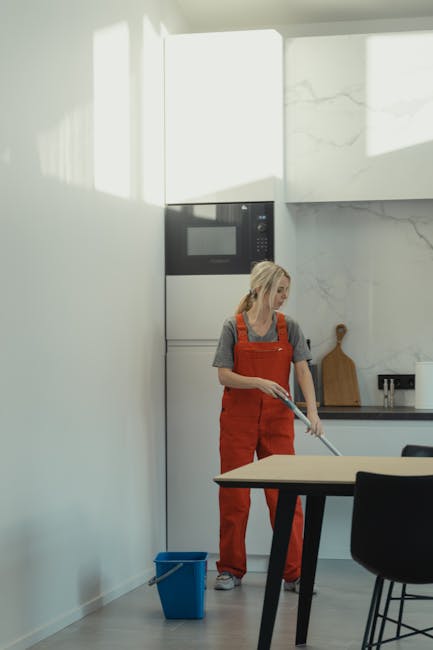 A woman in casual clothing and an orange apron is engaged in surface cleaning within a modern kitchen. She holds a mop or cleaning tool and is positioned near a white wall with a window that allows natural light to illuminate the space. The kitchen features a built-in microwave oven integrated into the cabinetry, a marble-effect splashback, and a black countertop with a wooden cutting board and a small set of utensils. On the floor, there is a blue cleaning bucket, indicating active cleaning or sanitisation. The surfaces, including the table and countertops, appear tidy and well-maintained, reflecting a thorough domestic cleaning process conducted by Hammersmith Cleaners as part of their same-day cleaning service in Hammersmith.