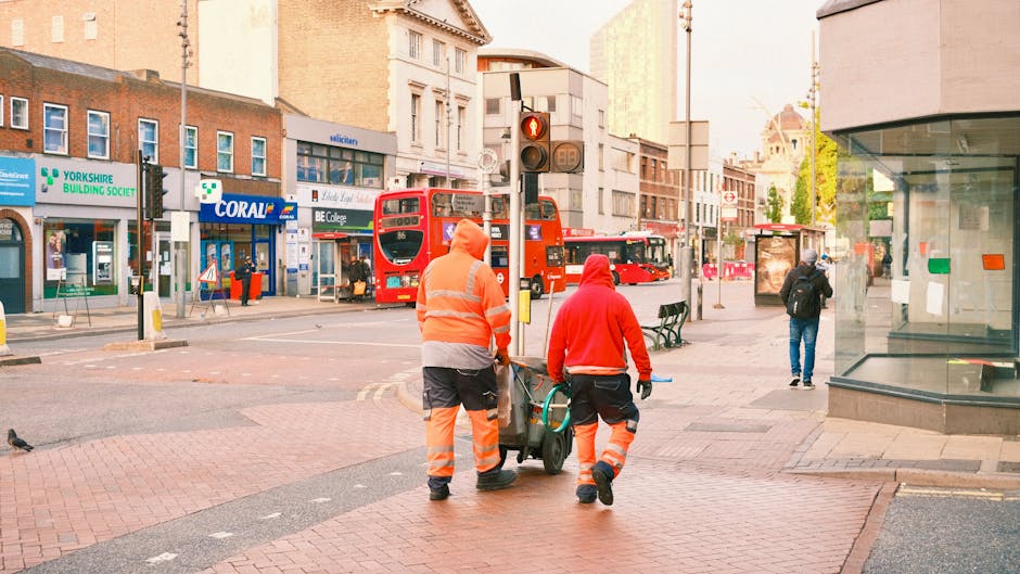 Two workers from Hammersmith Cleaners dressed in high-visibility clothing, including orange and red jackets with reflective stripes, are engaged in surface cleaning or maintenance activity on a city street. They are using a small trolley or cleaning cart, which holds cleaning supplies and tools. The scene is set on a paved pedestrian area with red brick and stone surfaces near a busy intersection. In the background, there are buildings with retail stores, including a Yorkshire Building Society branch, a Coral shop, and a college entrance. A red double-decker bus is visible at the tram stop, along with a few pedestrians walking or standing nearby. The street is well-lit with natural daylight, and the overall scene reflects urban cleanliness and maintenance efforts essential to city hygiene and public space upkeep, as provided by Hammersmith Cleaners.